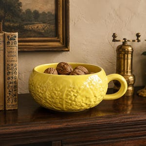 May include: A bright yellow ceramic bowl with a single handle and a textured, embossed surface pattern resembling cabbage leaves. The bowl is filled with several whole brown walnuts, showcasing a rounded shape and a glossy, smooth finish.