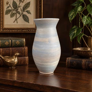 May include: A ceramic vase with a flared rim and a tapered body, featuring a horizontal brushstroke pattern in soft shades of light blue, cream, and white. The surface has a matte finish with visible texture from the application of the paint.