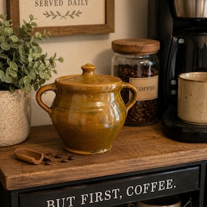 May include: A rustic, glazed ceramic coffee pot with a lid and two handles sits on a wooden shelf. The pot is a warm, earthy yellow-brown color. A sign above reads "COFFEE SERVED DAILY." The phrase "BUT FIRST, COFFEE" is visible on the shelf below.
