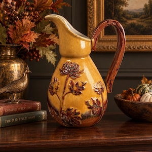 May include: A glazed ceramic pitcher in a warm mustard yellow hue, featuring a prominent relief design of dark brown and rust-colored chrysanthemum flowers and foliage. The vessel has a curved handle and a wide pouring spout.