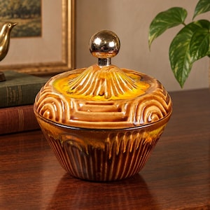 May include: A ceramic lidded bowl featuring a glossy brown and yellow drip glaze finish. The bowl has a textured, ribbed exterior with geometric patterns and a removable lid topped with a smooth, reflective gold-toned spherical knob handle.