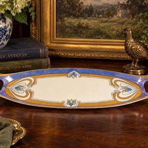 May include: An oval ceramic serving tray featuring a central cream-colored panel bordered by gold and blue bands. The tray includes integrated handles on each end and is decorated with small blue floral motifs at the top, bottom, and sides.