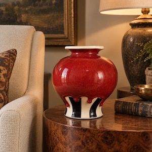 May include: A ceramic vase with a glossy red upper section that features a drip-like pattern over a base decorated with vertical black and white stripes. The vase has a wide, rounded body and a smooth, white-rimmed opening at the top.