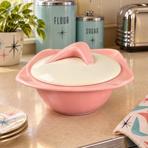 May include: A pink ceramic serving bowl with a wide, flared rim and a matching off-white lid. The lid features a prominent, curved, boomerang-shaped handle in pink. The bowl has a smooth, glossy finish and a rounded base, presenting a distinct mid-century aesthetic.