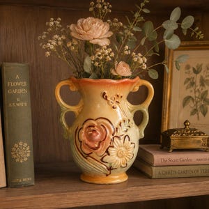 May include: A ceramic vase with two handles featuring a gradient of orange, yellow, and green tones. The surface is decorated with embossed floral patterns, including a large central rose and smaller daisy-like flowers, holding a bouquet of flowers.