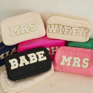 May include: A collection of rectangular zippered pouches in various colors including cream, black, hot pink, and light pink. Each pouch features large, white, varsity-style block letters with gold glitter trim spelling out words like MRS, WIFEY, and BABE.