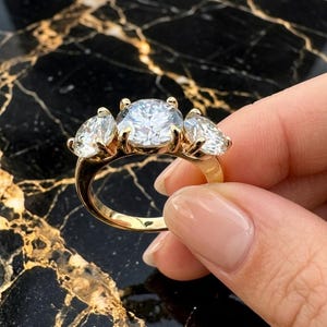 Peut inclure: Une bague en or avec une grande pierre centrale taille ronde entourée de deux pierres plus petites taille ovale. Les trois pierres sont maintenues par des griffes en or sur un anneau en or poli, formant un design à trois pierres.