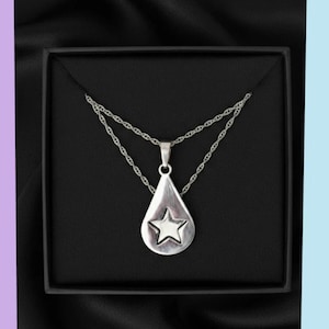May include: A silver-toned necklace featuring a teardrop-shaped pendant with a centered five-pointed star design. The pendant hangs from a delicate twisted chain, presented inside a square black box with a smooth, dark interior lining.