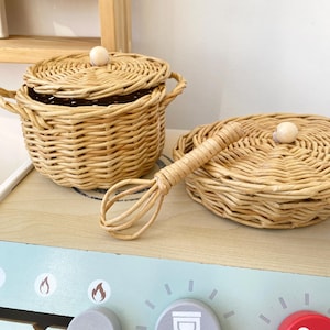 Può includere: Un set di giocattoli da cucina in rattan intrecciato che include una pentola con coperchio, una padella piatta con coperchio e una frusta. Gli oggetti sono marrone chiaro e presentano piccoli pomelli in legno sui coperchi, su un fornello giocattolo.