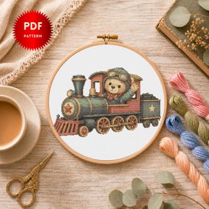 Peut inclure: Motif de point de croix d'un ours en peluche conduisant un train vintage, encadré dans un cercle à broder en bois. Le train est orné de rouge, de vert et d'or. L'image comprend une tasse de thé, des ciseaux et du fil à broder.
