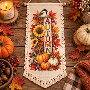 May include: A rectangular wall hanging featuring a cross-stitch design of autumn elements including a bird, sunflowers, pumpkins, and red berries. The word AUTUMN is vertically embroidered in red letters. The fabric has a cream lace border and fringe.