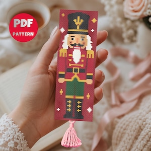 May include: A rectangular bookmark featuring a pixelated nutcracker design on a red background with a pink tassel at the bottom. The nutcracker wears a black hat, red jacket with gold accents, and green trousers. The text PDF PATTERN is visible.