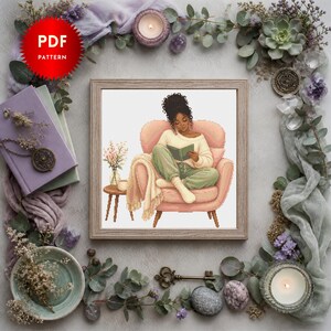 May include: A digital PDF cross stitch pattern featuring an illustration of a person with dark curly hair sitting in a pink armchair, reading a book while wearing a cream sweater and green trousers, with a small side table and flowers nearby.