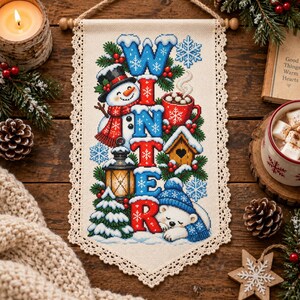 Peut inclure: Une tenture murale rectangulaire avec un motif au point de croix sur tissu crème et une bordure en dentelle. Le mot WINTER est écrit en grandes lettres bleues et rouges, entouré d'un bonhomme de neige, d'un mug de cacao et d'un ours polaire.