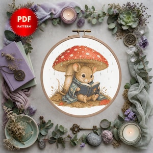 May include: A cross-stitch pattern featuring a brown mouse sitting under a large red mushroom with white spots, wearing a blue plaid blanket and reading a small book. The design is displayed in a round wooden embroidery hoop on a white background.