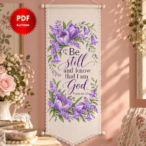 Könnte beinhalten: Ein vertikaler Wandbehang in Kreuzstichoptik mit violetten Tulpen und Fliederblüten, die den Text "Be still and know that I am God. Psalm 46:10" umrahmen. Der cremefarbene Stoff hat eine Spitzenborte und hängt an einer Holzstange.
