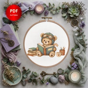 May include: A cross-stitch pattern of a teddy bear in a pilot's hat and scarf, reading a book, set in a wooden embroidery hoop. The image includes books, pencils, and a coffee cup. The text "PDF PATTERN" is in a red circle.