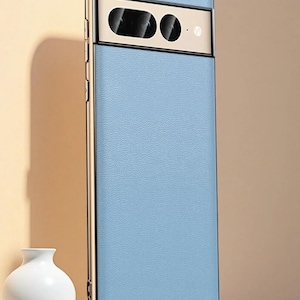 Könnte beinhalten: Eine hellblaue Smartphone-Hülle mit strukturierter Oberfläche und einem metallisch goldenen Rahmen. Die Hülle verfügt über einen horizontalen Kameraausschnitt mit goldenem Rand, der zwei schwarze Linsen und einen kleinen Blitz zeigt.