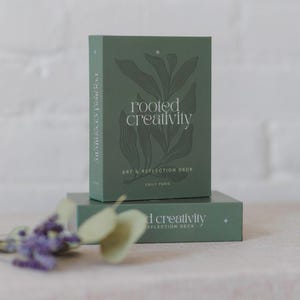May include: A sage green rectangular box set titled Rooted Creativity Art & Reflection Deck by Emily Paris, featuring a dark green botanical line art illustration on the front. A matching box base sits underneath with the same text and star icon.