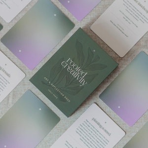 Könnte beinhalten: Ein Kartendeck mit dem Titel Rooted Creativity Art & Reflection Deck von Emily Paris, bestehend aus einer dunkelgrünen Schachtel mit botanischer Strichzeichnung. Mehrere Karten liegen verstreut und zeigen sanfte lila und grüne Farbverläufe.