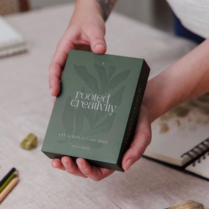 May include: A rectangular deck of cards in a dark olive green box. The front features a subtle, darker green botanical leaf illustration, with the white text "rooted creativity" and "ART & REFLECTION DECK" printed below, and "EMILY PARIS" at the bottom.