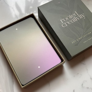 May include: A deck of cards with a gradient finish transitioning from muted green to soft purple, featuring small star icons. The cards are housed in a rectangular olive green box with the text "rooted creativity" and "ART & REFLECTION" printed on the lid.