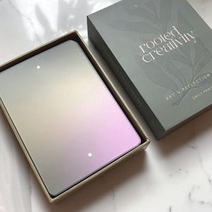May include: A deck of rectangular cards with a smooth gradient finish transitioning from muted green to soft purple, featuring small white star icons. The cards are housed in a sage green box with the text rooted creativity ART & REFLECTION EMILY PARIS.
