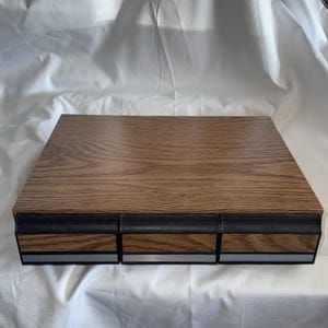 May include: A rectangular storage unit featuring a wood grain finish on the top surface and three front-facing drawers. Each drawer has a black textured handle, a matching wood grain panel, and a thin silver horizontal trim along the bottom edge.