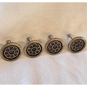 May include: Four circular metal knobs featuring a raised flower design with a dark center and a metallic gold-toned rim. Each knob has a short metal post extending from the back, designed for attachment to furniture or cabinetry surfaces.
