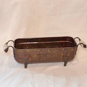 May include: Rectangular, antique-style metal planter with a distressed, copper-toned finish. Features decorative handles with white and blue ceramic accents and small feet. A vintage-inspired container for plants or decor.