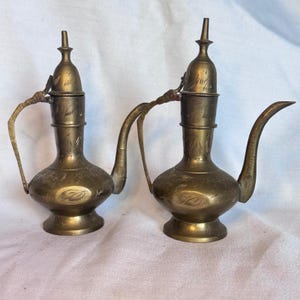 May include: Two brass ewers featuring a bulbous base, a slender neck, and a pointed lid. Each vessel has a curved handle and a long, tapered spout. The metal surface is decorated with etched leaf patterns and shows a weathered patina.