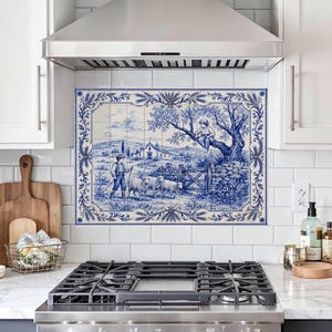 Puede incluir: Un mural de azulejos de cerámica decorativo con una escena pastoral en azul y blanco de un pastor con ovejas y una persona sentada en un árbol. El mural está enmarcado por un borde botánico detallado instalado sobre una estufa de cocina.