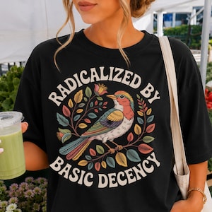 Op de afbeelding: Een zwart t-shirt met korte mouwen en een kleurrijke afbeelding van een vogel op een tak, omringd door bladeren en bloemen. De tekst RADICALIZED BY BASIC DECENCY staat in een cirkelvormig wit lettertype rondom de centrale illustratie.