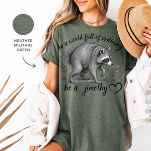 Peut inclure: Un t-shirt vert militaire chin&eacute; avec une illustration graphique noire repr&eacute;sentant un raton laveur. Le texte indique "In a world full of ordinary be a jimothy" avec un petit c&oelig;ur. Le motif inclut des &eacute;l&eacute;ments botaniques.