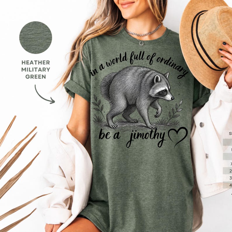 Peut inclure: Un t-shirt vert militaire chin&eacute; avec une illustration graphique noire repr&eacute;sentant un raton laveur. Le texte indique "In a world full of ordinary be a jimothy" avec un petit c&oelig;ur. Le motif inclut des &eacute;l&eacute;ments botaniques.