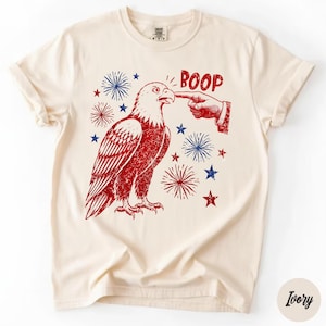 Könnte beinhalten: Ein elfenbeinfarbenes T-Shirt mit einer rot-blauen Grafik eines Weißkopfseeadlers, dessen Schnabel von einem Finger berührt wird. Das Design enthält den Schriftzug BOOP in roten Buchstaben, umgeben von roten und blauen Feuerwerkskörpern und Sternen.