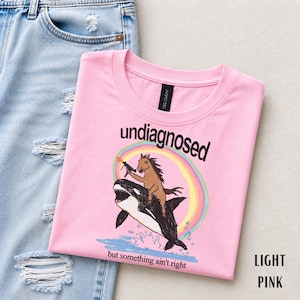 Può includere: Una t-shirt rosa chiaro con la grafica di un cavallo marrone che cavalca un'orca sotto un arcobaleno. Il cavallo tiene in mano una piccola arma da fuoco. Il testo "undiagnosed" è sopra la grafica e "but something ain't right" sotto.