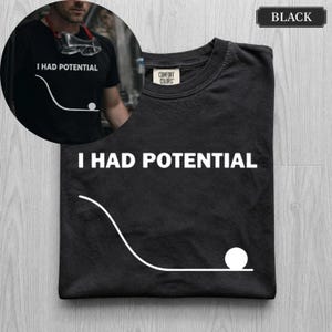 Puede incluir: Una camiseta negra de manga corta con el texto blanco I HAD POTENTIAL y un gráfico de una bola en la parte inferior de una pendiente curva. La camiseta tiene una etiqueta Comfort Colors en el cuello interior y se muestra plana con una imagen circular.