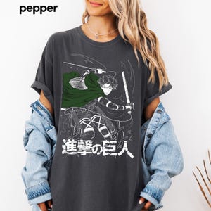 Op de afbeelding: Een antracietgrijs t-shirt met een grafische print van een anime-personage met een groene cape die een zwaard vasthoudt. Het ontwerp bevat witte lijntekeningen en Japanse karakters. Het t-shirt heeft korte mouwen en een ronde hals, gedragen met een lichtblauw spijkerjack.