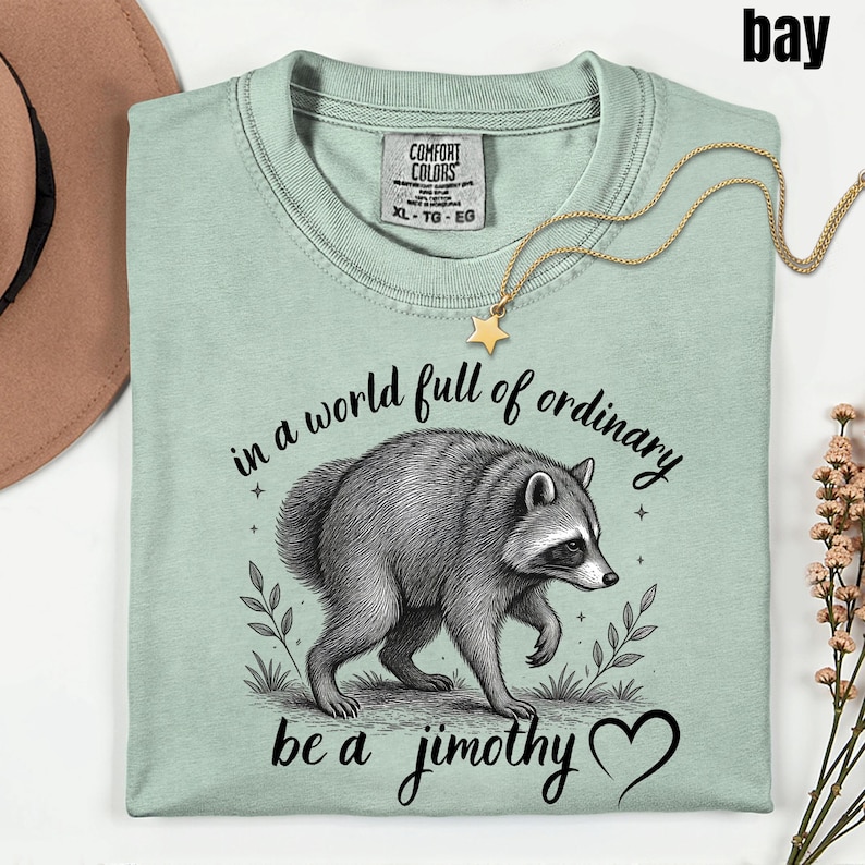 Peut inclure: Un t-shirt vert clair avec une illustration en noir et blanc d'un raton laveur. Le texte indique "in a world full of ordinary be a jimothy" avec un symbole de c&oelig;ur. Le t-shirt pr&eacute;sente une &eacute;tiquette Comfort Colors au col rond.
