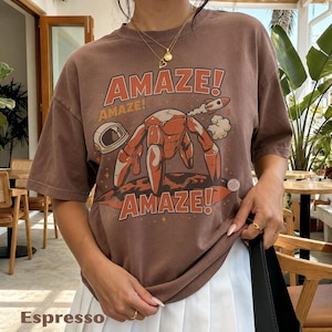 May include: A brown short-sleeve t-shirt featuring a graphic print of a crab in a space suit with a rocket and an astronaut helmet. The text AMAZE! is printed in large orange letters above and below the illustration. The shirt has a crew neckline.