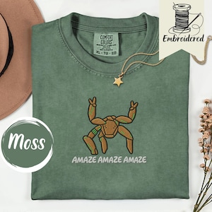 May include: A moss green t-shirt featuring an embroidered brown spider-like creature with green accents, making peace signs with two legs. The text "AMAZE AMAZE AMAZE" is embroidered below the creature. The shirt has a Comfort Colors label.