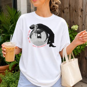 May include: A white short-sleeve t-shirt featuring a black and white graphic of a person draped over a large disco ball. The text "KISS ALL THE TIME" and "DISCO OCCASIONALLY" is printed in red in a circular pattern around the central graphic.