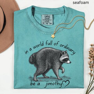 Peut inclure: Un t-shirt &agrave; manches courtes vert mousse avec une illustration de raton laveur et le texte "in a world full of ordinary be a jimothy" accompagn&eacute; d'un c&oelig;ur. Le t-shirt pr&eacute;sente une &eacute;tiquette Comfort Colors au col et une encolure ronde.