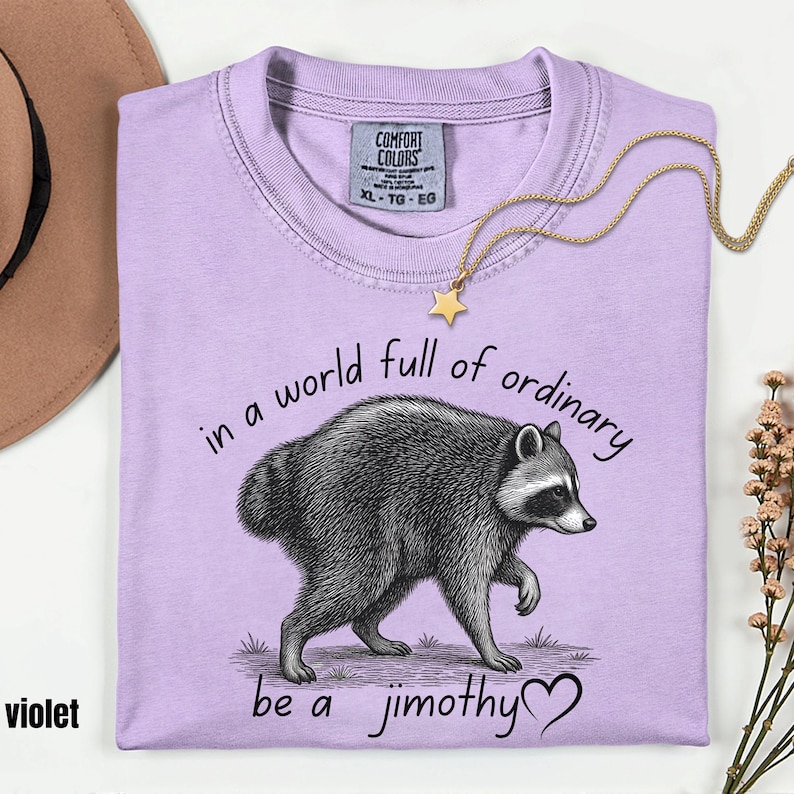 Peut inclure: Un t-shirt violet clair avec une illustration en noir et blanc d'un raton laveur. Le texte indique "in a world full of ordinary be a jimothy" avec un symbole de c&oelig;ur. L'&eacute;tiquette Comfort Colors indique la taille XL, TG, EG.