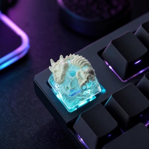 May include: A custom mechanical keyboard keycap featuring a white, sculpted skeletal dragon design atop a translucent, ice-like base. The keycap is illuminated from beneath by a bright cyan light, contrasting with the surrounding black keys.