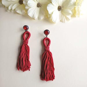 Puede incluir: Un par de pendientes largos de borlas rojas con un pendiente de piedra preciosa roja circular en la parte superior. Las borlas están hechas de múltiples hilos rojos, asegurados por una pequeña banda de cuentas sobre la franja principal.