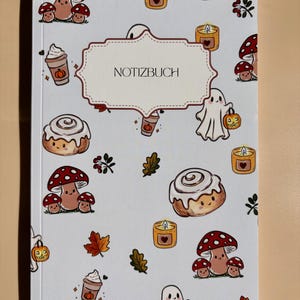 Könnte beinhalten: Ein weißes Notizbuch mit einem wiederkehrenden Muster aus herbstlichen Illustrationen, darunter rote Pilze, Zimtschnecken, Geister mit Kürbissen, Kerzen und Kaffeebechern. In der Mitte befindet sich ein dekoratives Etikett mit dem Text NOTIZBUCH.