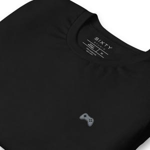 May include: Black t-shirt featuring a small, embroidered grey video game controller. The shirt has a crew neck and a label that reads "SIXTY STUDIO" and "Made in Nicaragua". The fabric is 100% combed ring spun cotton.