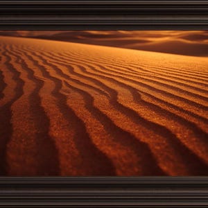 May include: A framed print depicting a desert landscape. The image shows rippled sand dunes in shades of orange and brown, illuminated by warm sunlight. The frame is dark brown.
